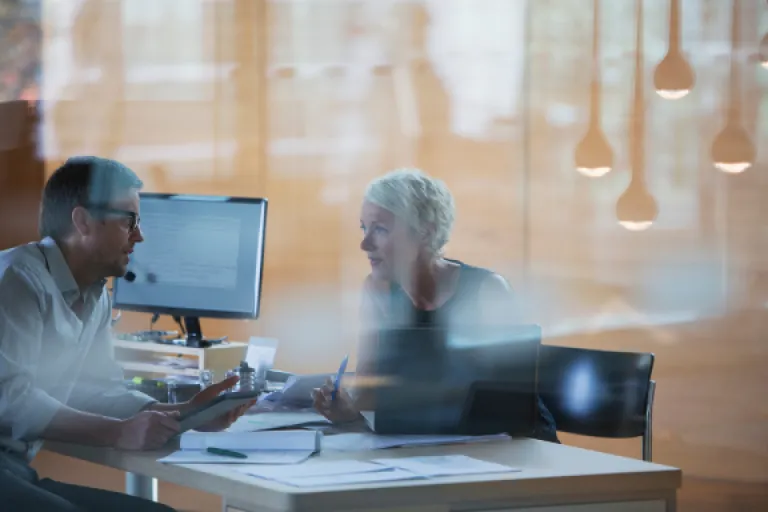 Two professionals having a discussion across a desk. The man is holding a notebook and the woman is holding a pen.