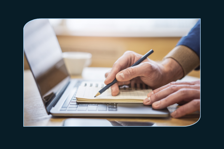 A close-up of a person's hands taking notes in a small notepad while working on a laptop. One hand holds a pen, and the other rests on the keyboard. A coffee cup sits in the blurred background. The person is wearing a blue sweater with a beige cuff.